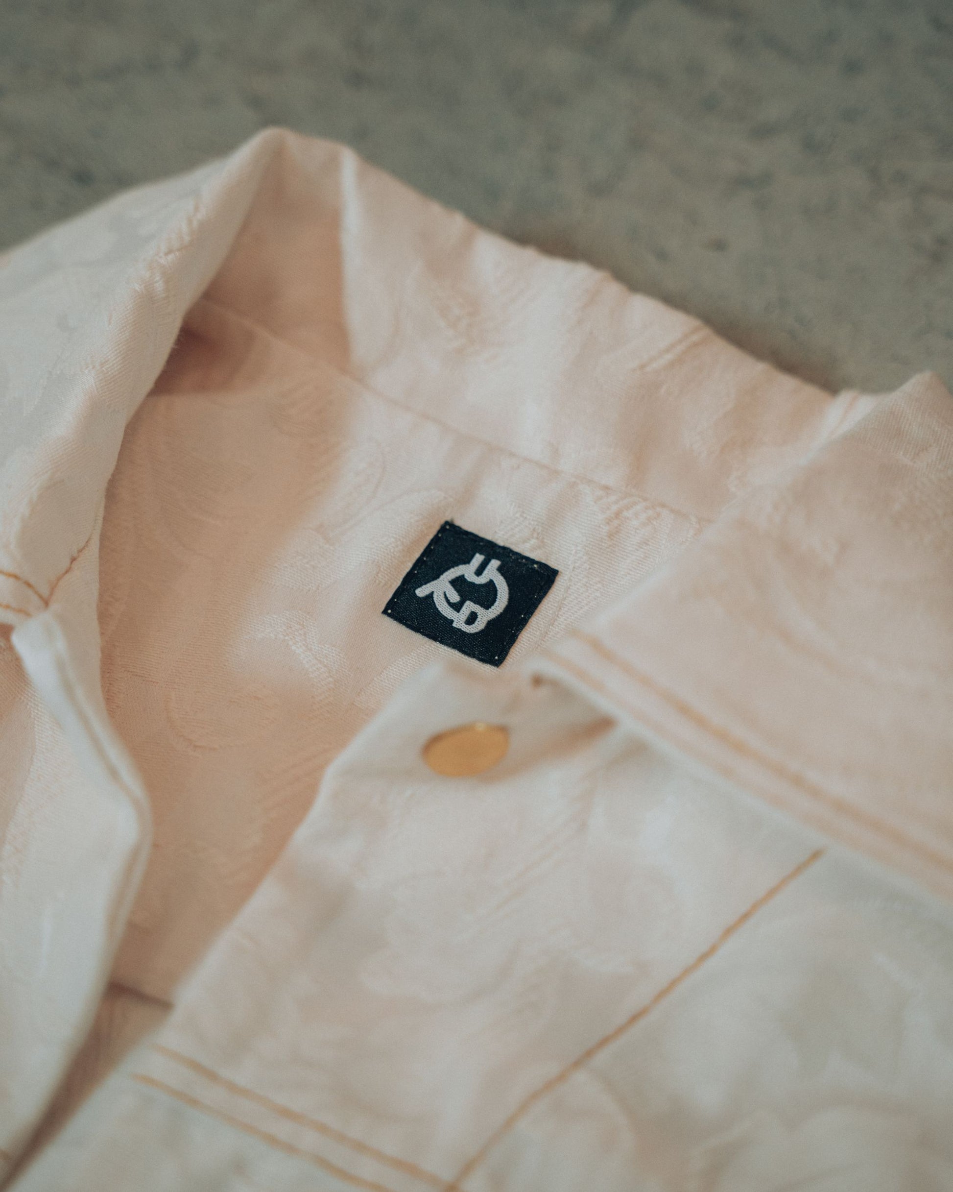 Close-up of a cream jacket with gold buttons and top stitching on a blurred background. Visible Uni Design brand logo.