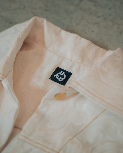 Close-up of a cream jacket with gold buttons and top stitching on a blurred background. Visible Uni Design brand logo.