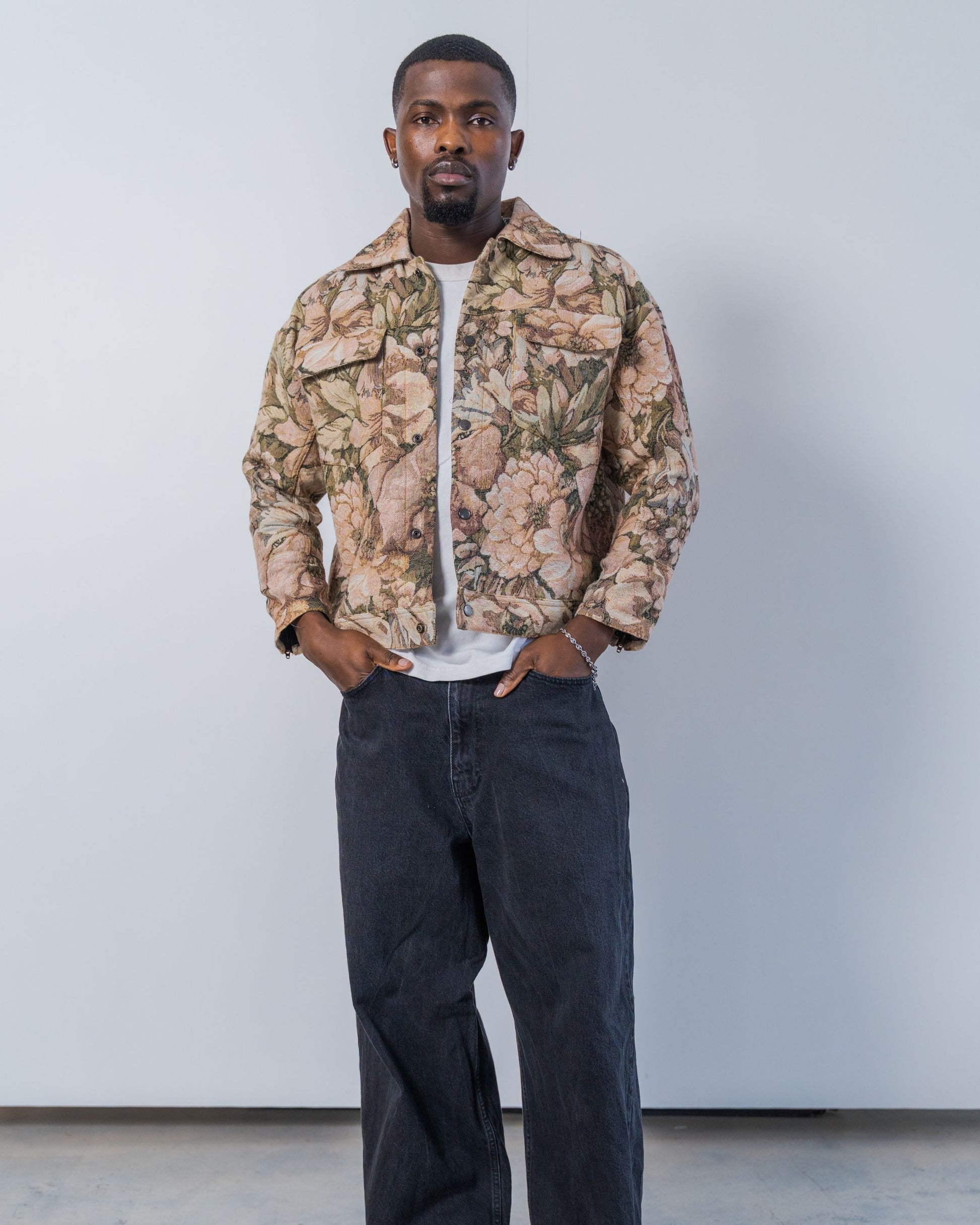 Man wearing a floral patterned jacket and dark pants against a plain background. The jacket is pink and brown with a jacquard floral print. It is sustainable and unisex. 