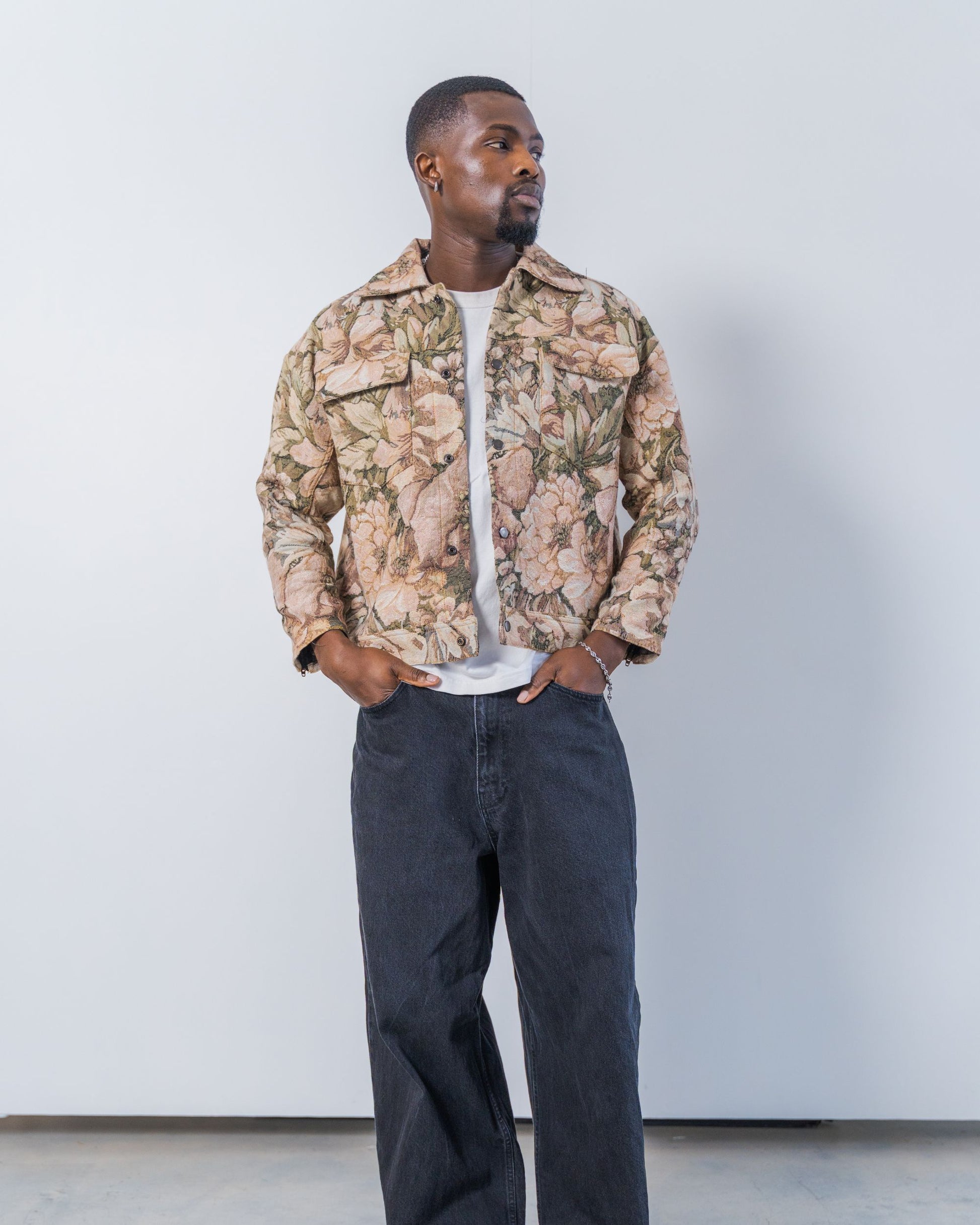 Man wearing a jacket and dark jeans against a plain background. The jacket is pink and brown with a jacquard floral print. It is sustainable and unisex. 
