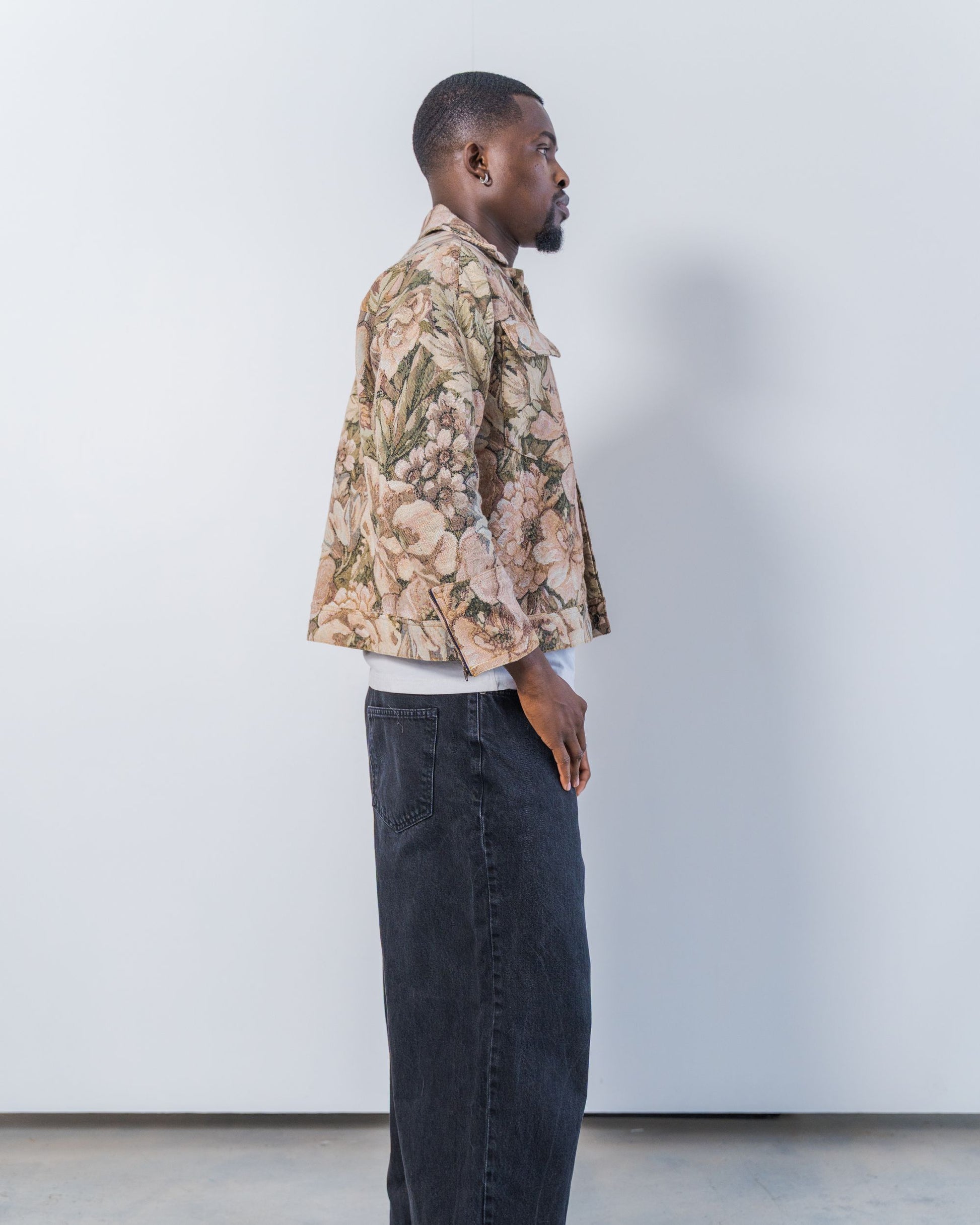 Man wearing a patterned jacket and dark jeans against a plain background. The jacket is pink and brown with a jacquard floral print. It is sustainable and unisex. 