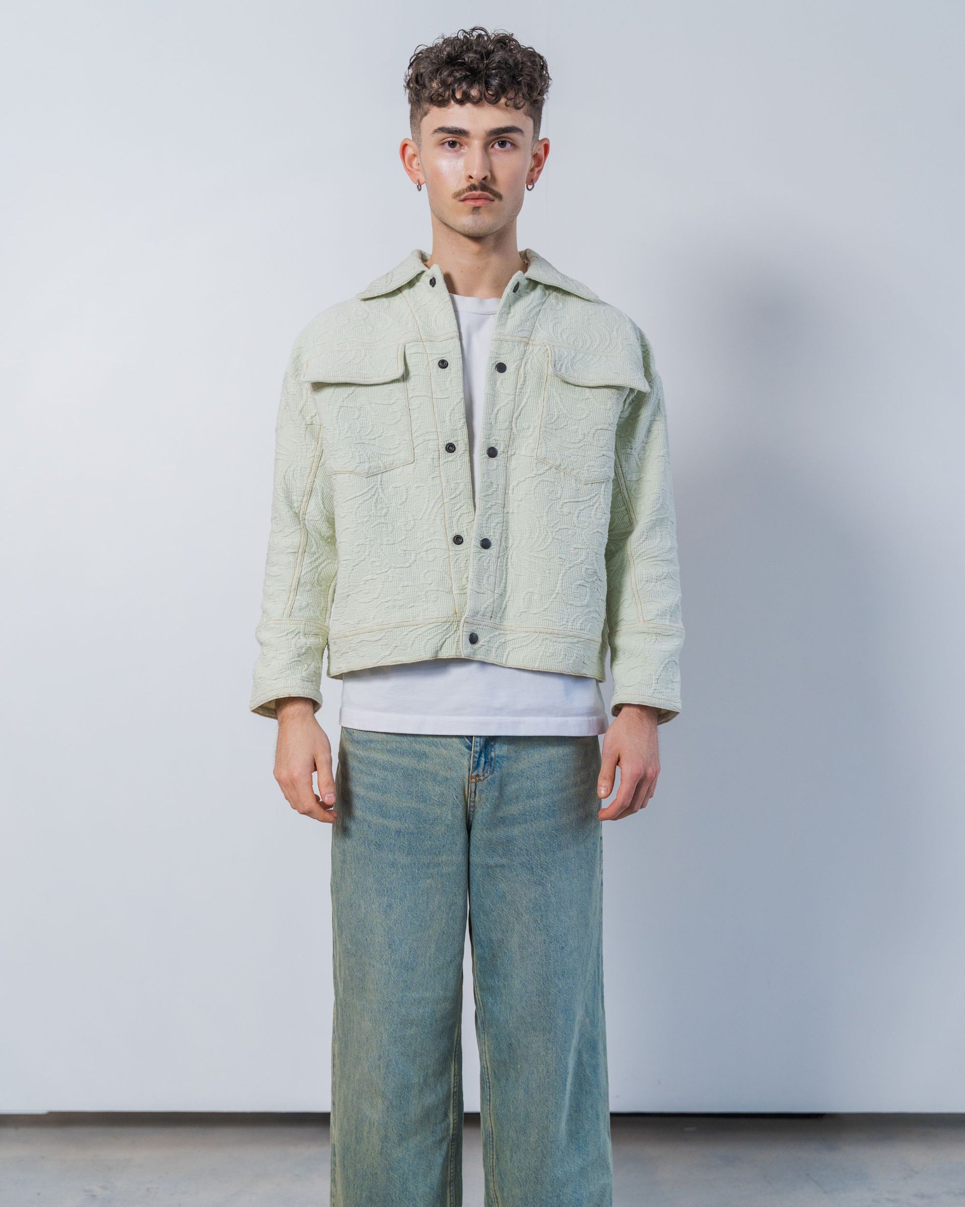 Man wearing a light green sage jacket and blue jeans against a plain background. the jacket is sustainable fashion made from upcycled vintage textiles.