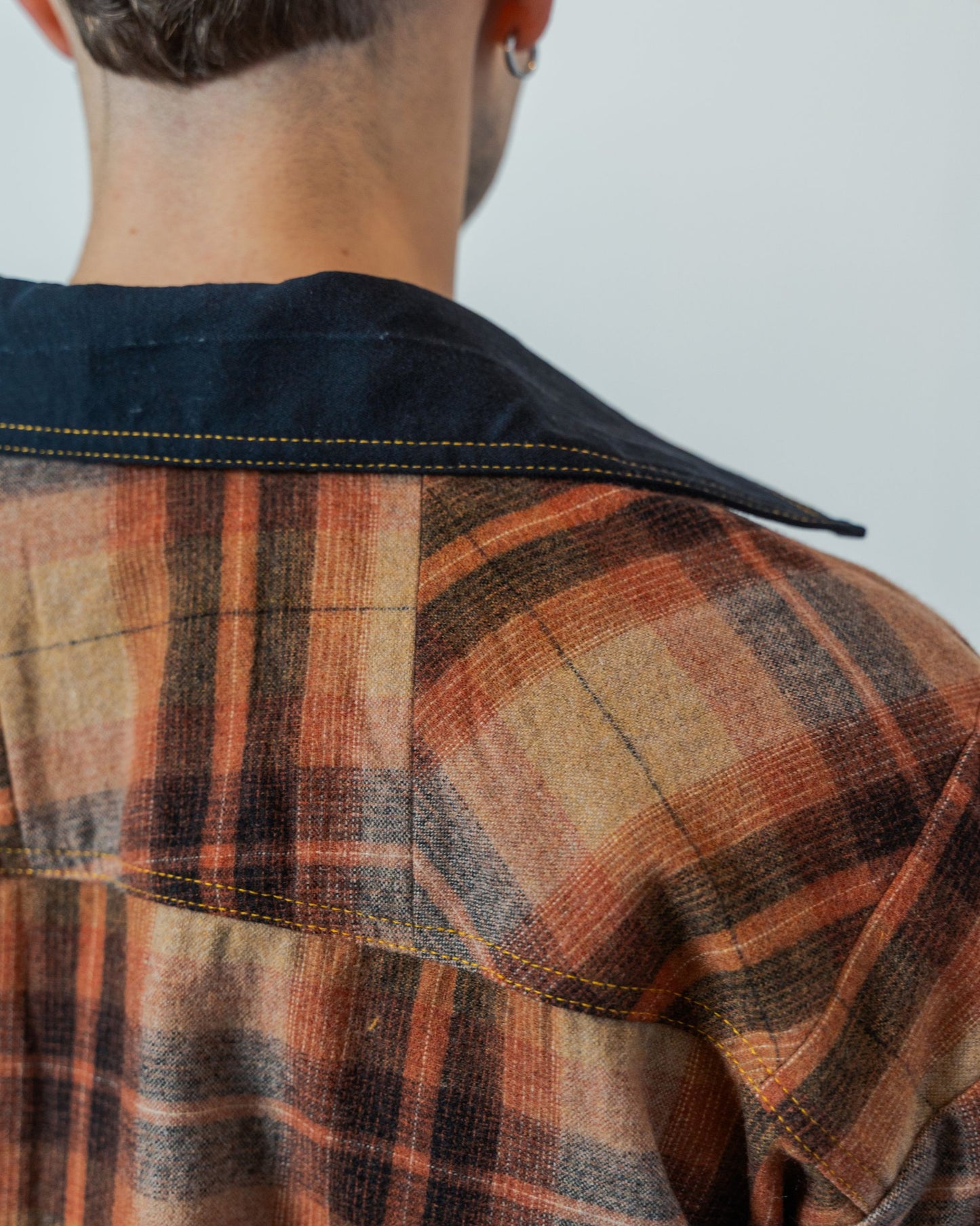 Person wearing a plaid shirt with a neutral background. Close up on the sustainable upcycled unisex jacket that shows black collar detail and patchwork on the back.