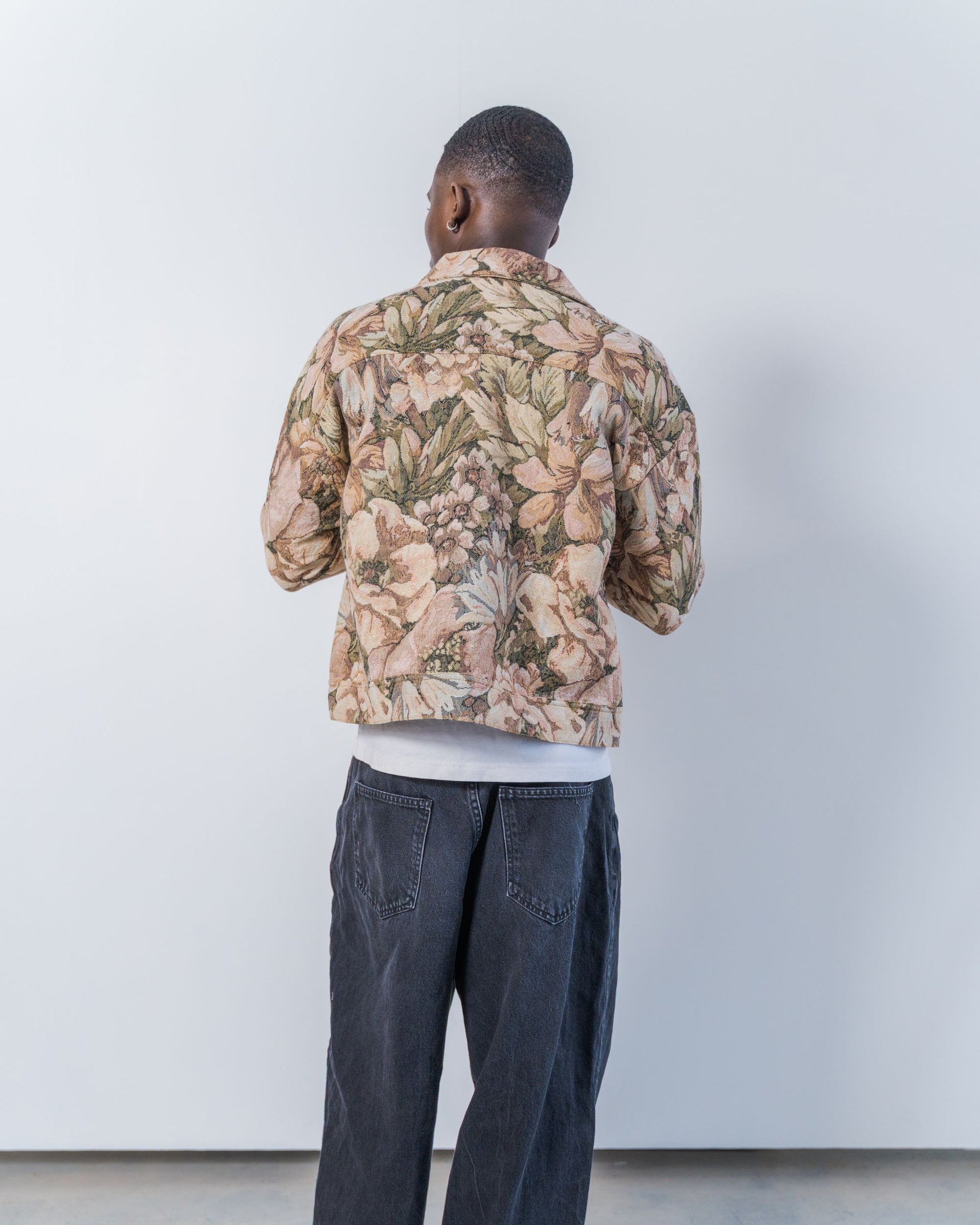 Person wearing a floral jacket and dark jeans against a white wall. The jacket is pink and brown with a jacquard floral print. It is sustainable and unisex. 