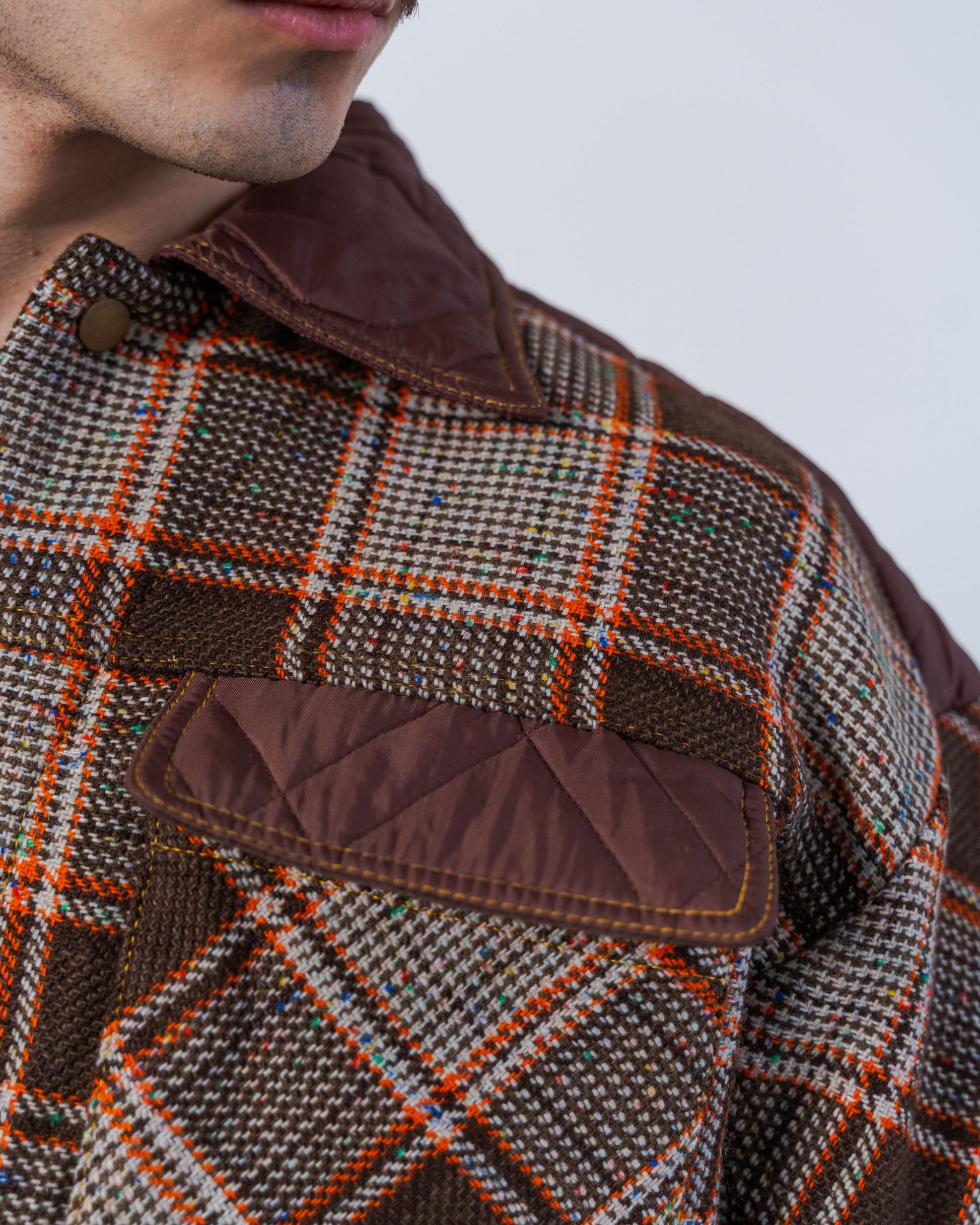 Close-up of a person wearing a checkered jacket with brown accents on a plain background. The jacket is unisex and made from sustainable upcycled materials. 
