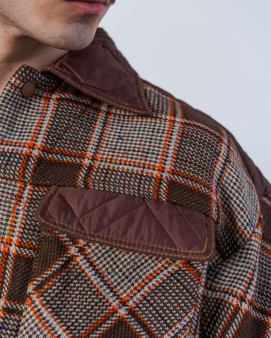 Close-up of a person wearing a checkered jacket with brown accents on a plain background. The jacket is unisex and made from sustainable upcycled materials. 