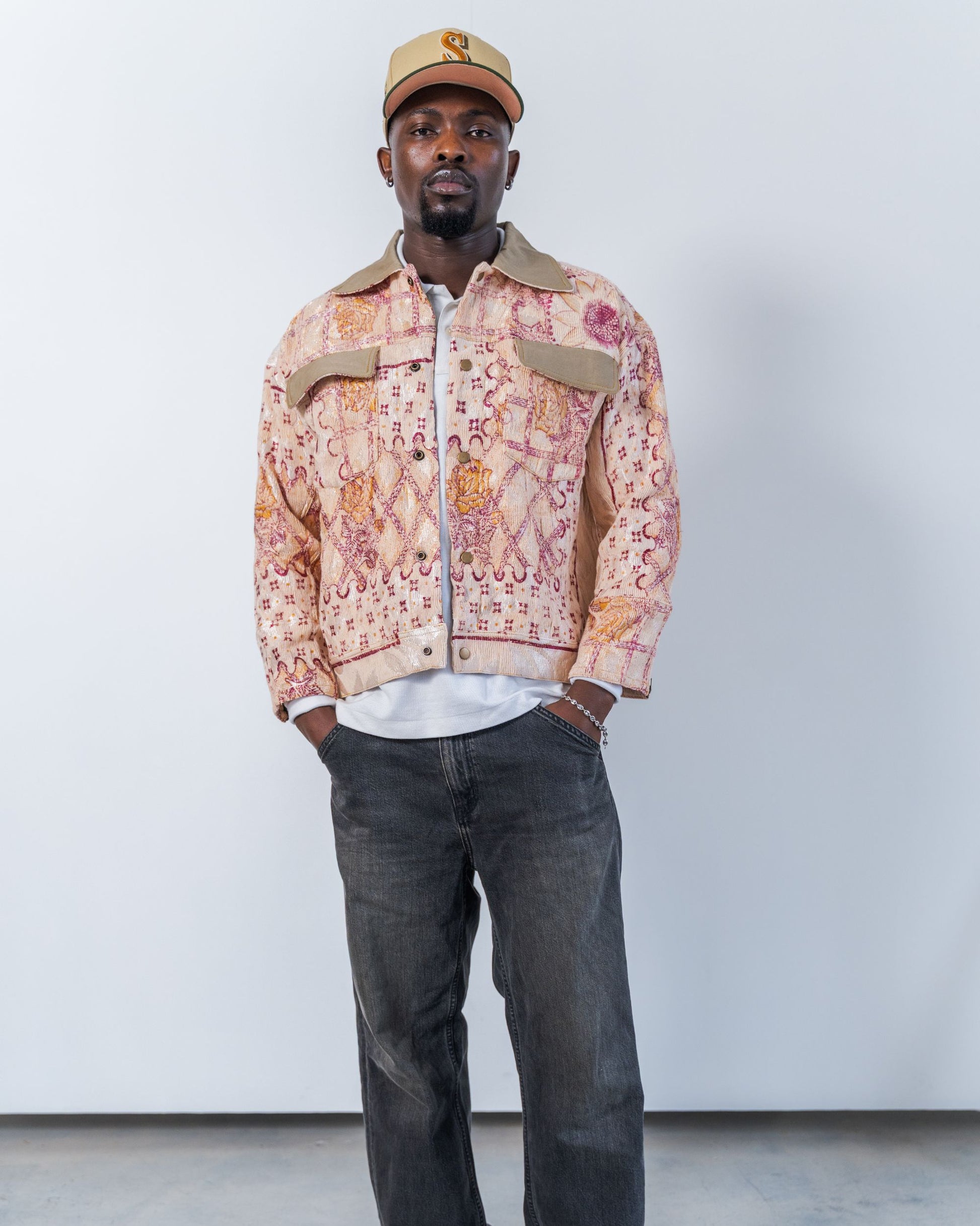 Man wearing a patterned jacket and cap against a plain background. The jacket is pink with floral print made from a vintage upcycled blanket. It is unisex, sustainable fashion.