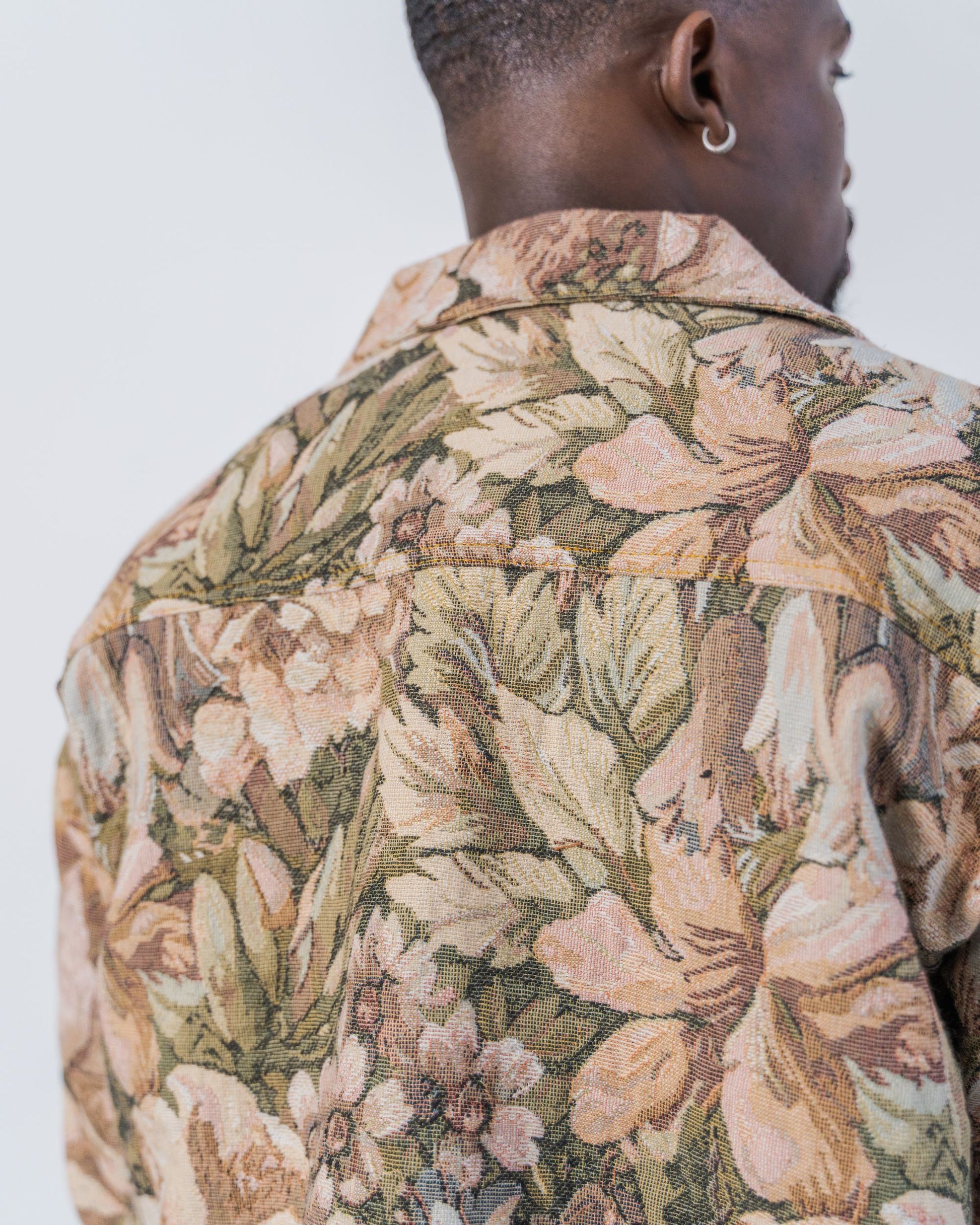 Person wearing a floral patterned shirt with a neutral background. The jacket is pink and brown with a jacquard floral print. It is sustainable and unisex. 