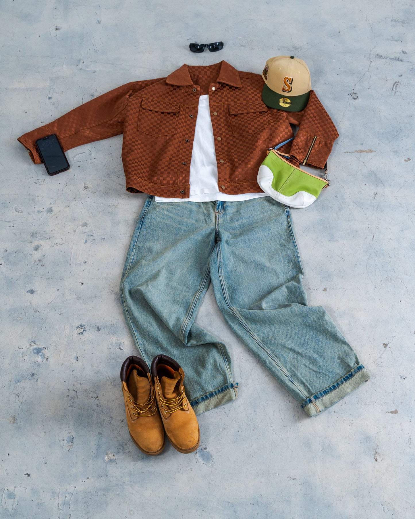 Brown jacket, blue jeans, tan boots, green bag, and cap on a light gray surface. Sustainable fashion outfit styled on the floor