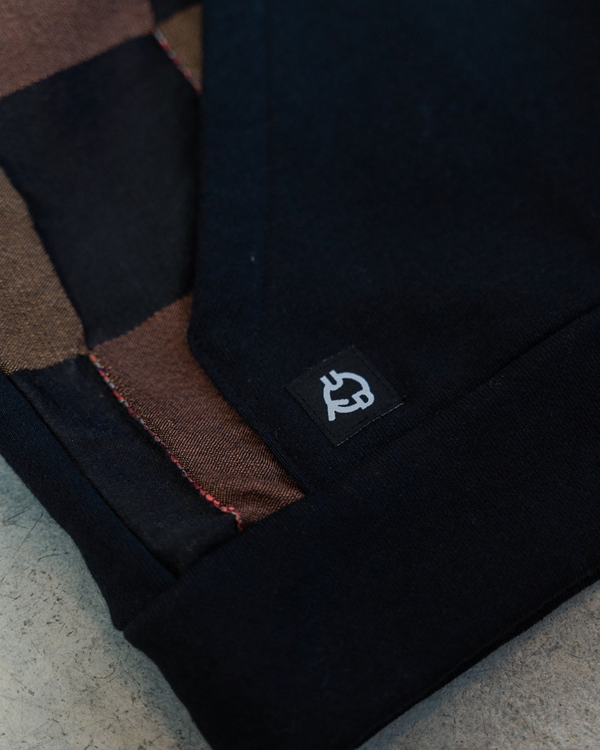 Close-up of a black hoodie pocket with Uni Design logo on a concrete floor. The close up shows the stitchwork on black and brown checkered fabric.